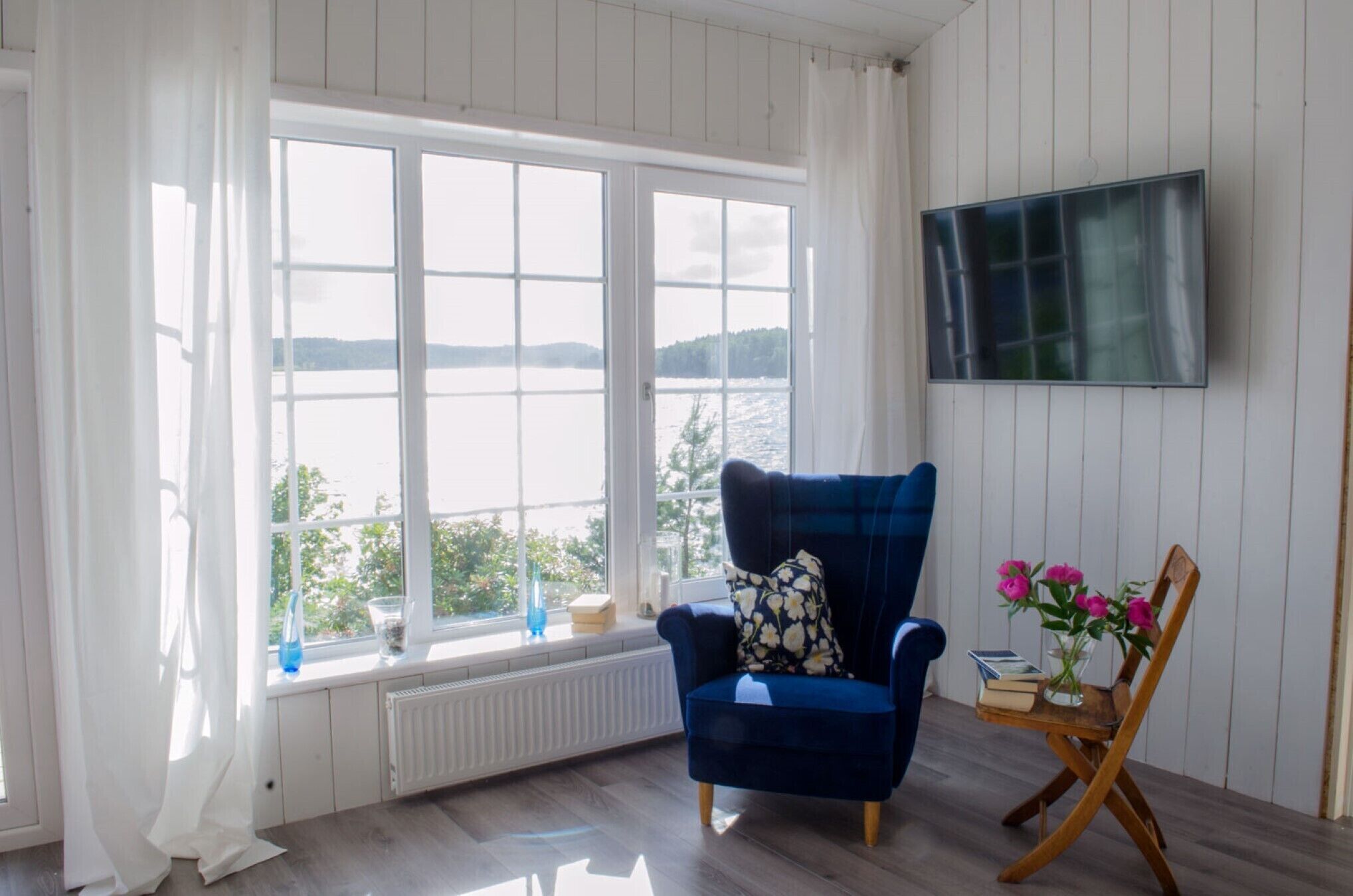 Ein Zimmer mit großem Fenster und Blick auf ein Gewässer, ein blauer Sessel mit floralem Kissen, ein Holzstuhl mit Blumenvase sowie ein wandmontierter Flachbildfernseher.