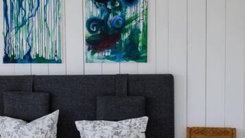 Ein Schlafzimmer mit einem Bett, zwei abstrakten Gemälden, einem grauen Kopfteil und einem hölzernen Stuhl mit einer weißen Lampe.