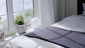 A bedroom with a large window overlooking a body of water, a bed with a dark headboard, and a small plant on the windowsill.