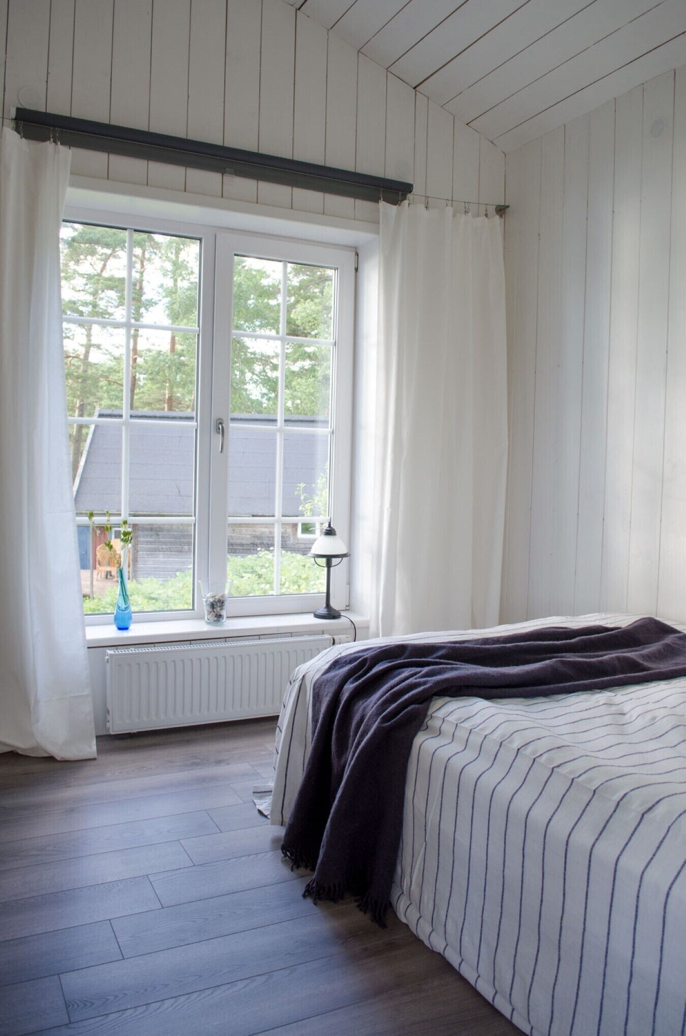 Quarto com uma janela grande, uma cama com roupa de cama listrada e um radiador abaixo da janela.