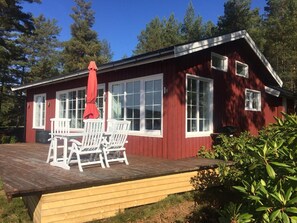 En rød hytte med en træterrasse, vinduer med hvide rammer og en rød parasol.