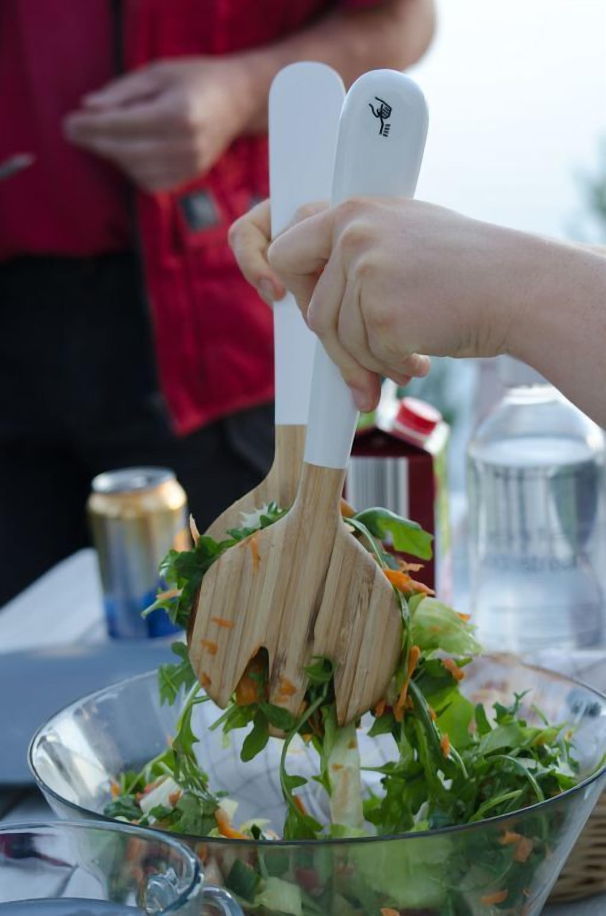 A person using wooden salad tongs to mix a salad in a clear glass bowl.