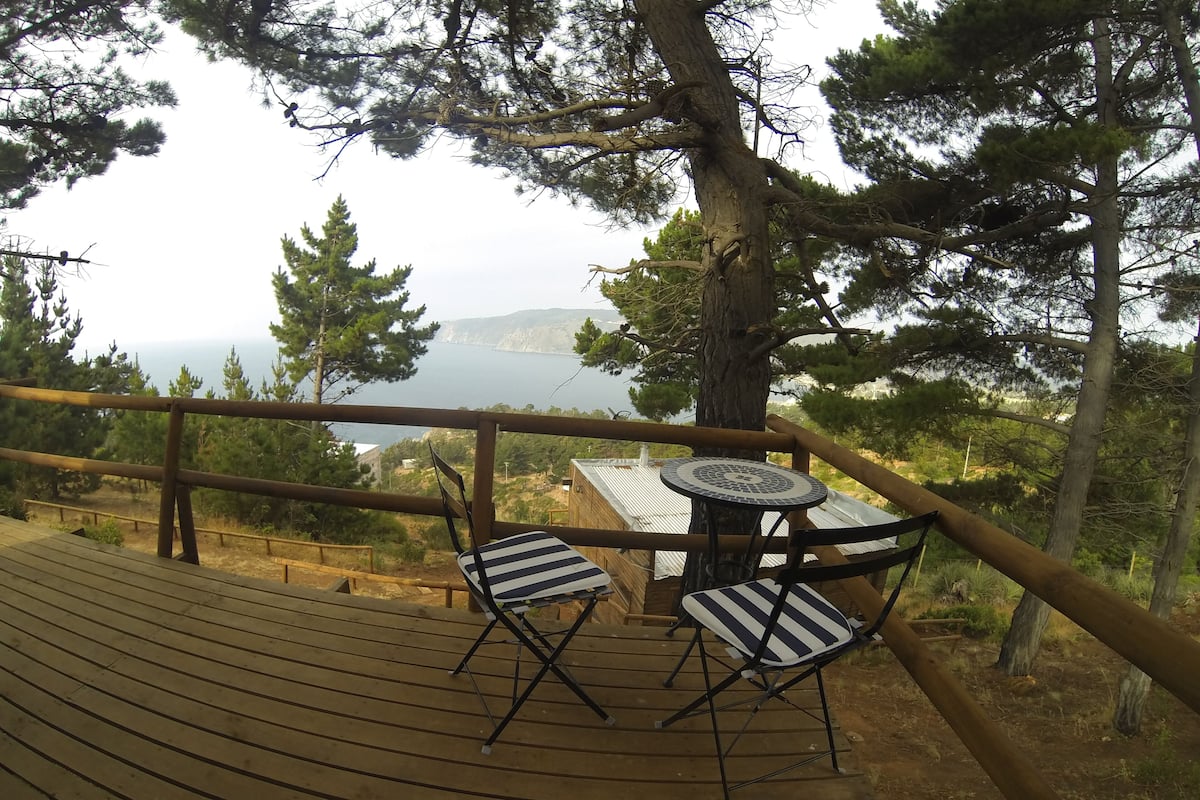 Una terraza de madera con una mesa y dos sillas, con vistas a un paisaje pintoresco de agua y montañas.