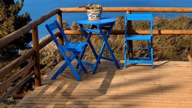 Terraza de madera con sillas azules y una mesa, con vista a un paisaje costero con acantilados.
