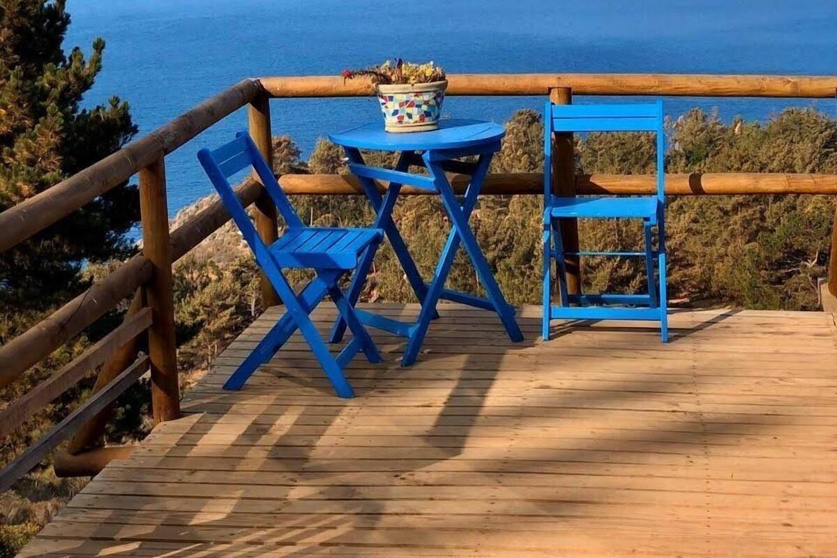 Terraza de madera con sillas azules y una mesa, con vista a un paisaje costero con acantilados.