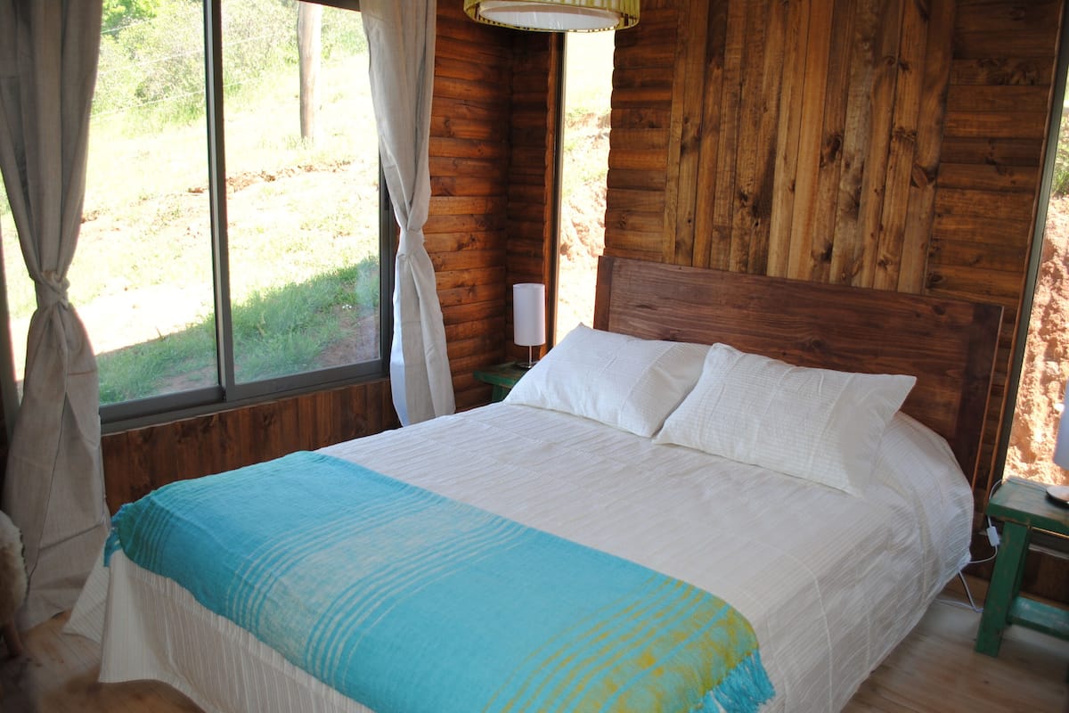 Un dormitorio de cabaña de madera con cama, cortinas y mesita de noche.