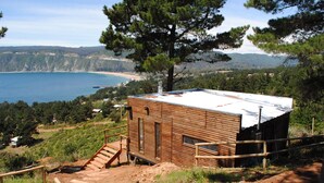 Una cabaña de madera con techo metálico, ubicada en una ladera con vistas a la costa.