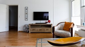 A living room with a wooden floor, a flat-screen TV on a wooden stand, a chair with a cushion, and a glass coffee table.
