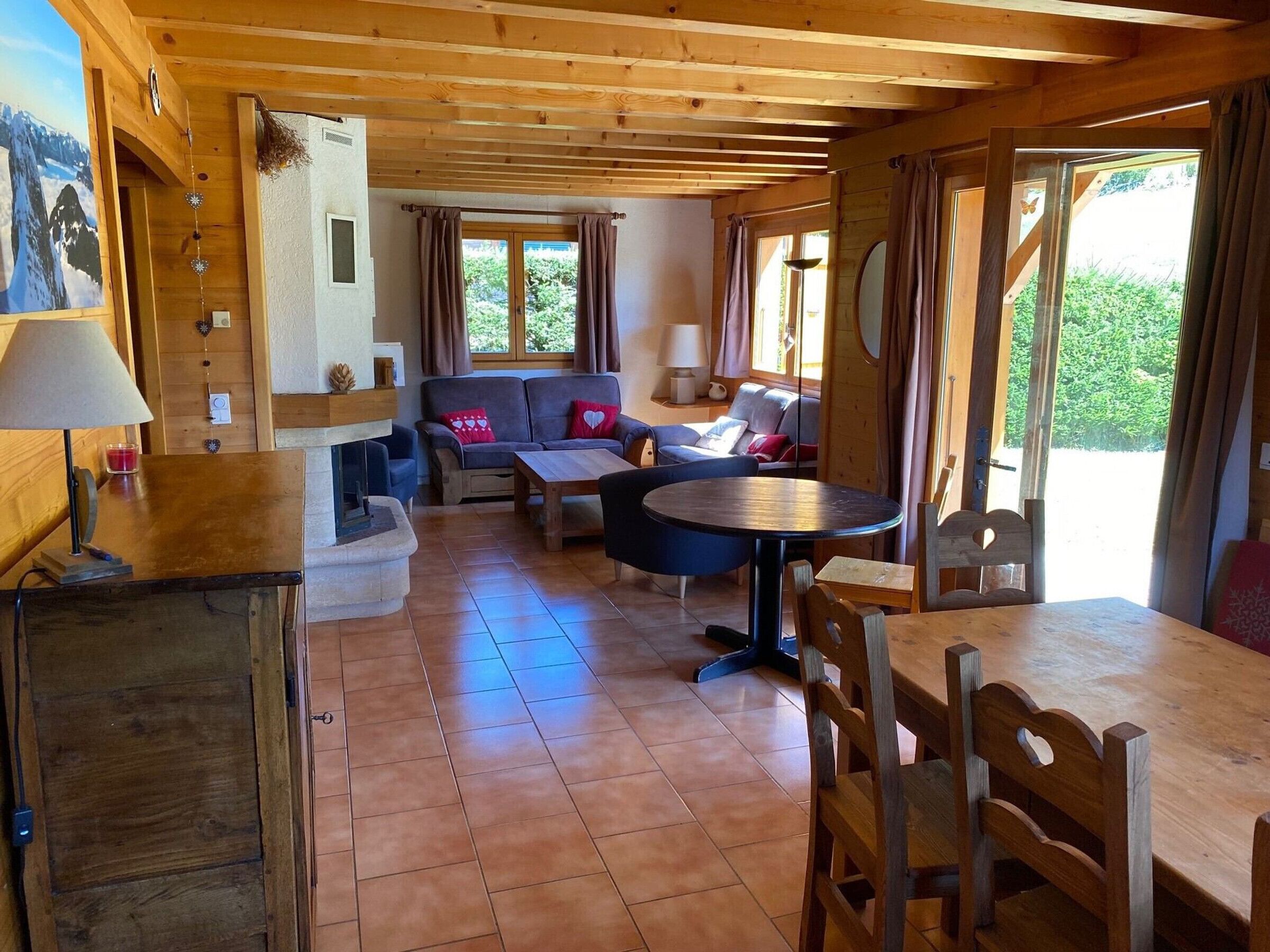 A cozy cabin interior with a fireplace, wooden furniture, and large windows offering a view of greenery.