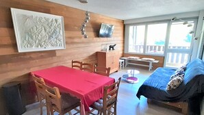 Um quarto aconchegante com parede de madeira, uma mesa de jantar com cadeiras, uma cama com cobertor azul, uma televisão fixada na parede e uma janela com vista para o exterior.