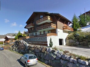 Una chaleta de varios pisos con balcones, un muro de piedra de contención y un coche estacionado al frente.