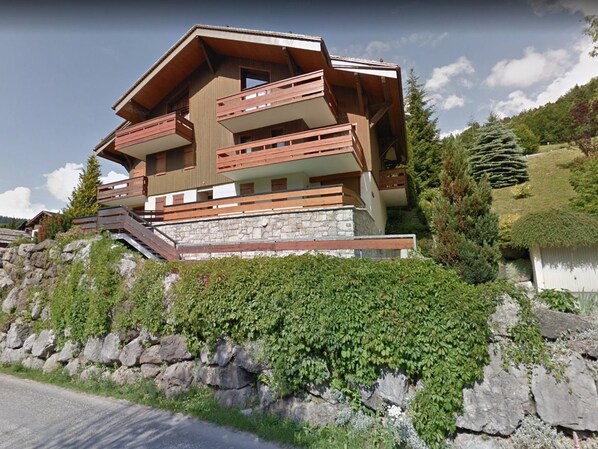 A modern multi-story chalet with wooden balconies, stone foundation, and lush greenery surrounding it.