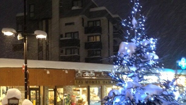 Área externa coberta de neve com um pinheiro enfeitado para o Natal, um edifício com letreiros iluminados e um poste de luz.