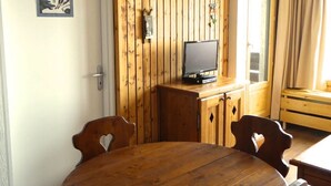 Um quarto com painéis de madeira, uma mesa redonda de madeira e duas cadeiras, uma televisão sobre um móvel de madeira, e uma janela com cortinas.