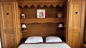 Quarto de madeira com cama, duas mesas de cabeceira e prateleira com objetos decorativos.