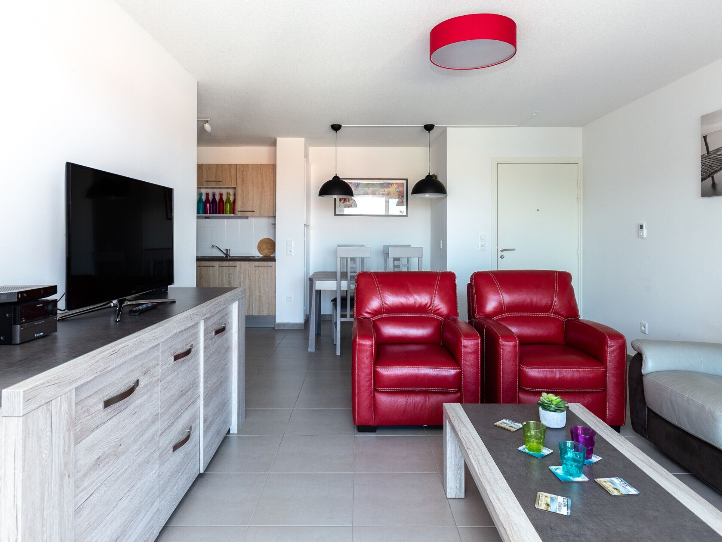 Un salon moderne avec un téléviseur, des fauteuils en cuir rouge, une table basse avec des boissons et, au fond, un coin repas.