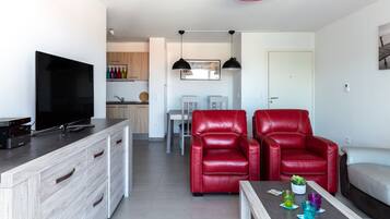 Un salon moderne avec un téléviseur, des fauteuils en cuir rouge, une table basse avec des boissons et, au fond, un coin repas.
