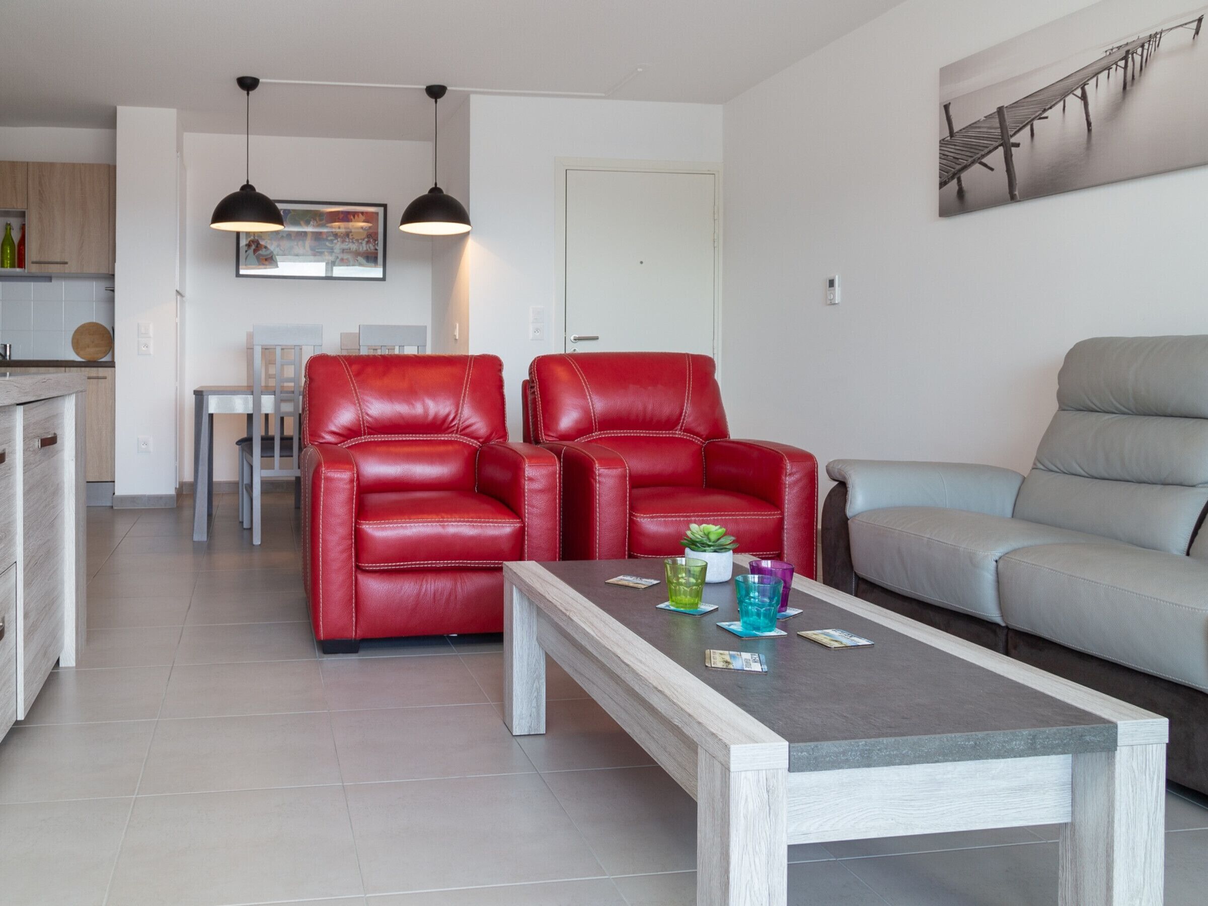 Un salon moderne avec un canapé gris, deux fauteuils en cuir rouge, une table basse en bois sur laquelle sont posés des magazines et des verres, et un coin repas à l’arrière-plan.