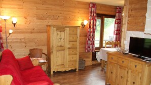 Un interior acogedor de cabaña con piso de madera, un sofá rojo, un mueble de madera con cajones, un televisor y una ventana con cortinas.