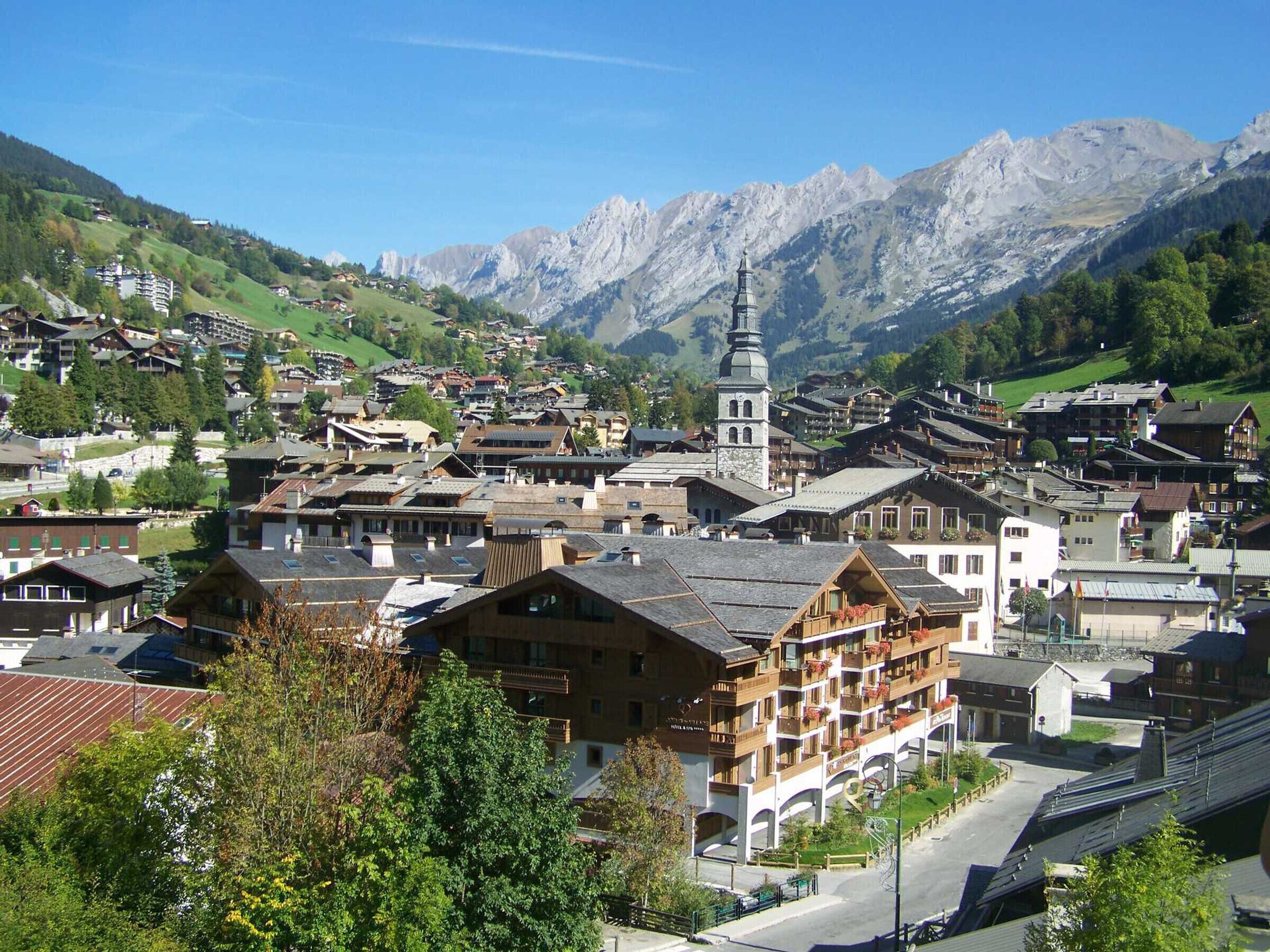 A traditional European village with historic buildings, a prominent church tower, and mountainous backdrop.