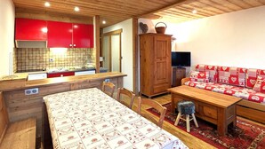 A cozy room with a wooden ceiling, a red kitchen area, a wooden dining table, and a sofa with heart-patterned cushions. - Cozy apt 4-6 pers balcony, ski-in/ski-out, covered parking (La Clusaz)
