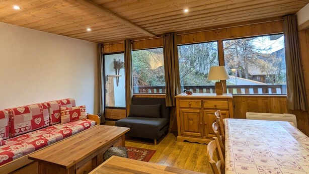 A cozy room with a wooden ceiling, a sofa with a heart-patterned blanket, a small table, a chair, a cabinet, and a window with a view of trees and a house. - Cozy apt 4-6 pers balcony, ski-in/ski-out, covered parking (La Clusaz)