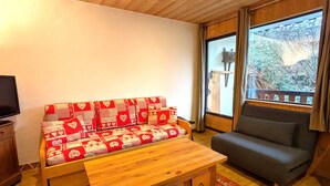 A cozy living room with a sofa covered in a red and white patterned blanket, a dark grey armchair, a wooden coffee table, and a large window with a view of trees. - Cozy apt 4-6 pers balcony, ski-in/ski-out, covered parking (La Clusaz)