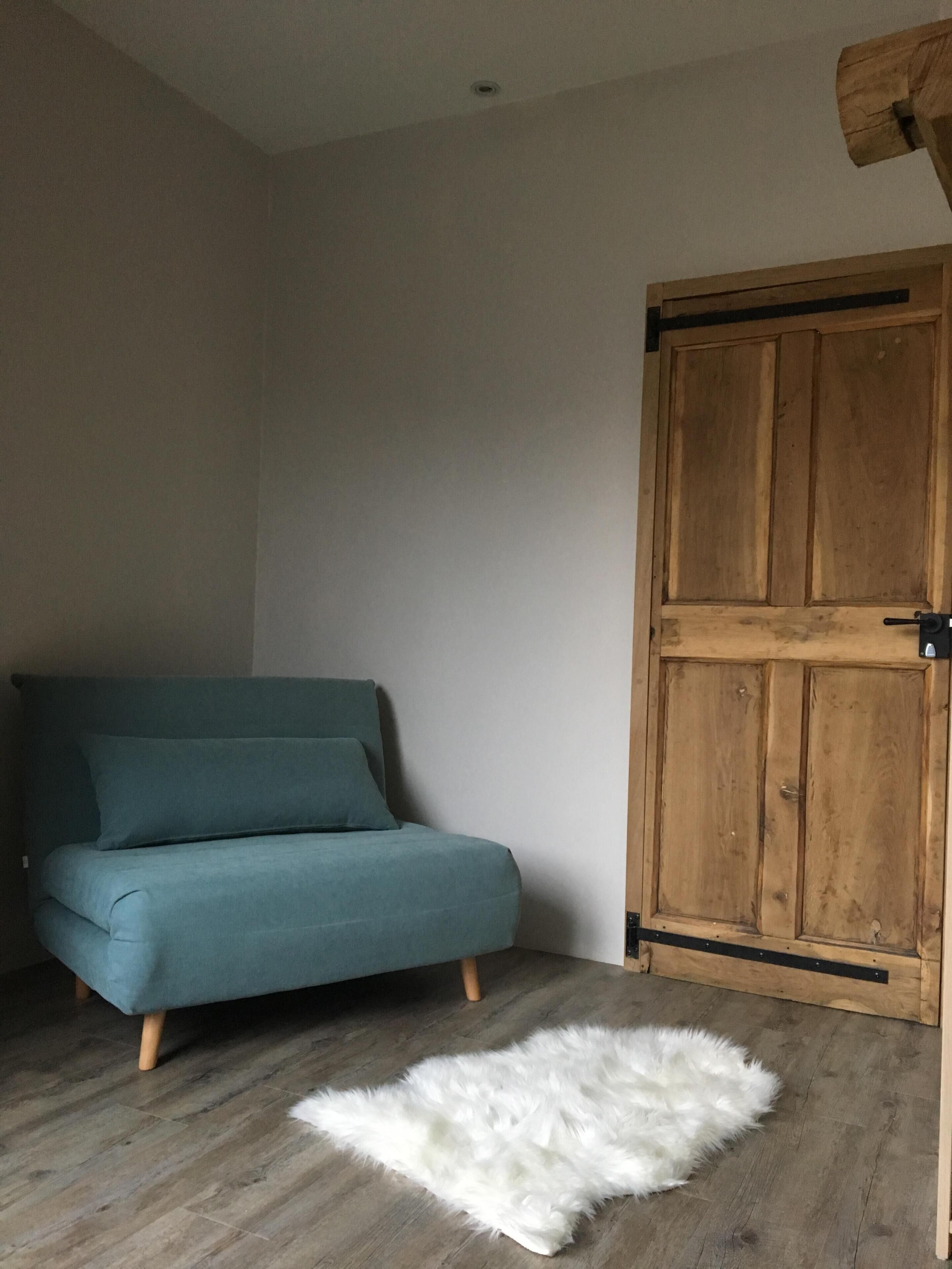 Ein Zimmer mit einem türkisfarbenen Sofa, einer Holztür und einem weißen, flauschigen Teppich auf Holzdielen.