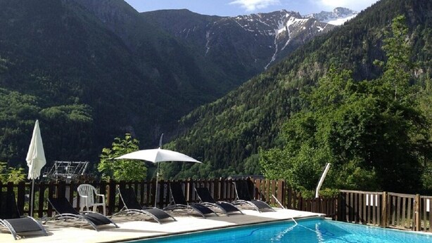 A swimming pool with lounge chairs and umbrellas, surrounded by a wooden fence, with a mountainous backdrop. - The 2 Alpes / Venosc, 10 pers. NEW with steam room, sauna, jacuzzi. (les deux alpes)