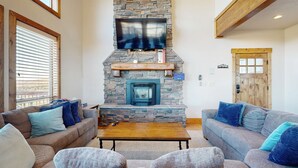 A living room with a stone fireplace, two sofas with blue cushions, a wooden coffee table, and a TV mounted above the fireplace.