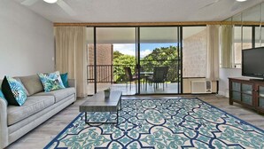 Un salón con sofá, una mesa de centro y un tapete estampado. Cuenta con un ventanal corredero que da a un patio exterior con sillas y un televisor.