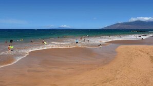 Una playa con gente y tablas de surf, cielo despejado y una montaña al fondo.