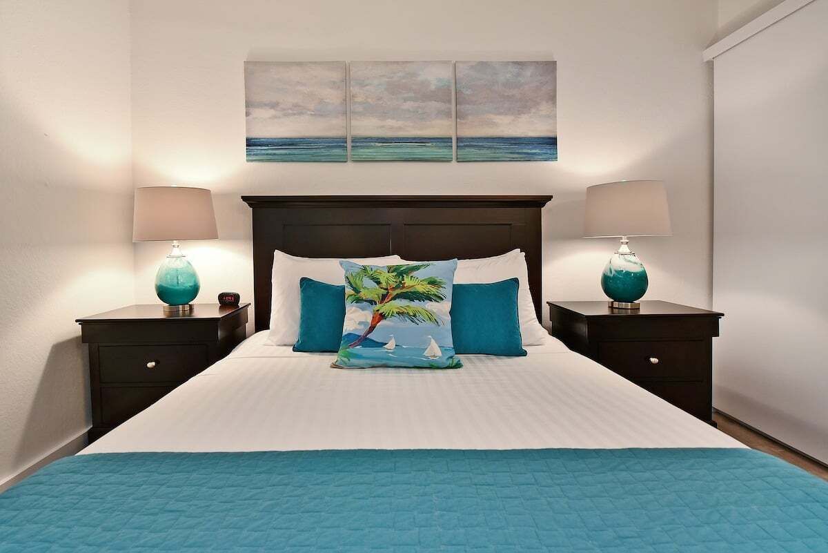Een net opgemaakt bed met een blauwe quilt, twee bijpassende nachtkastjes en een sierkussen met een tropisch thema.