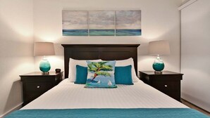 Una cama bien hecha con un edredón azul, dos mesitas de noche a juego y una almohada con motivos tropicales.