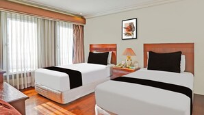 Habitación de hotel con dos camas, una mesita de noche con lámpara, un cuadro enmarcado en la pared y una ventana con cortinas.