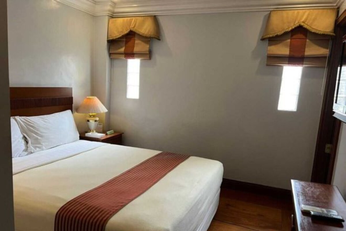 A hotel room with a bed, nightstand, lamp, and two windows with curtains.