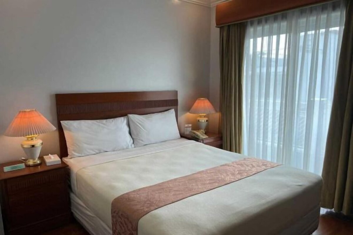 Habitación de hotel con una cama grande, dos lámparas de noche, una mesita de noche y una ventana con cortinas.