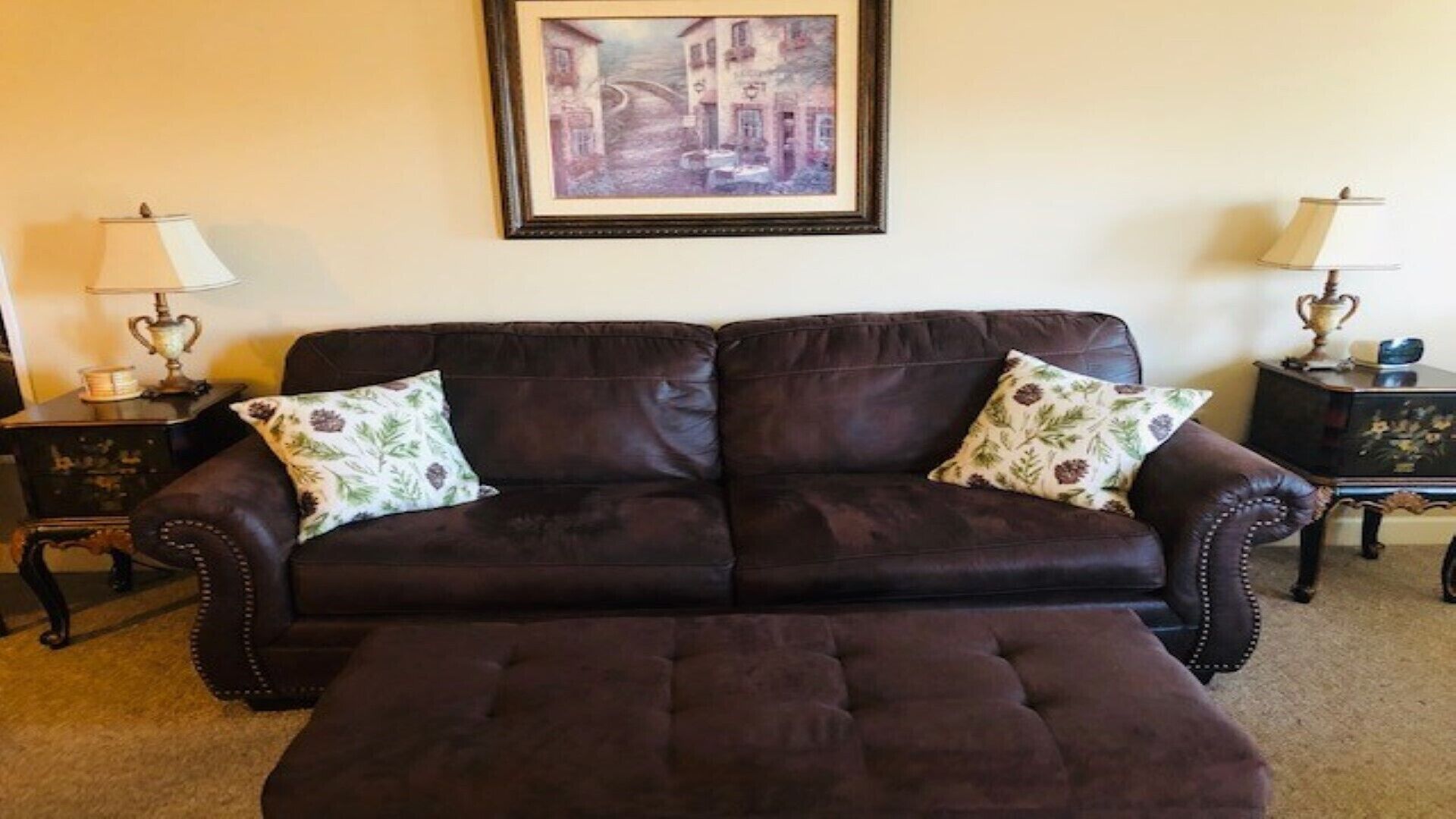 A dark brown leather sofa with floral cushions, flanked by two small side tables with lamps, against a plain wall with a framed painting above.