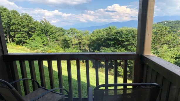 Eine Holzdielen-Terrasse mit zwei Stühlen, die auf eine grüne Fläche und eine bewaldete Landschaft mit Bergen im Hintergrund blickt.