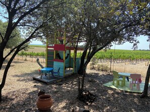 Un espace extérieur aménagé dans un style rustique, avec une table et des chaises en bois, une structure de jeu colorée et, en arrière-plan, un vignoble.