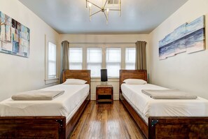Une chambre avec deux lits en bois, une table de chevet avec une lampe, une grande fenêtre avec des stores et des œuvres d’art accrochées aux murs.