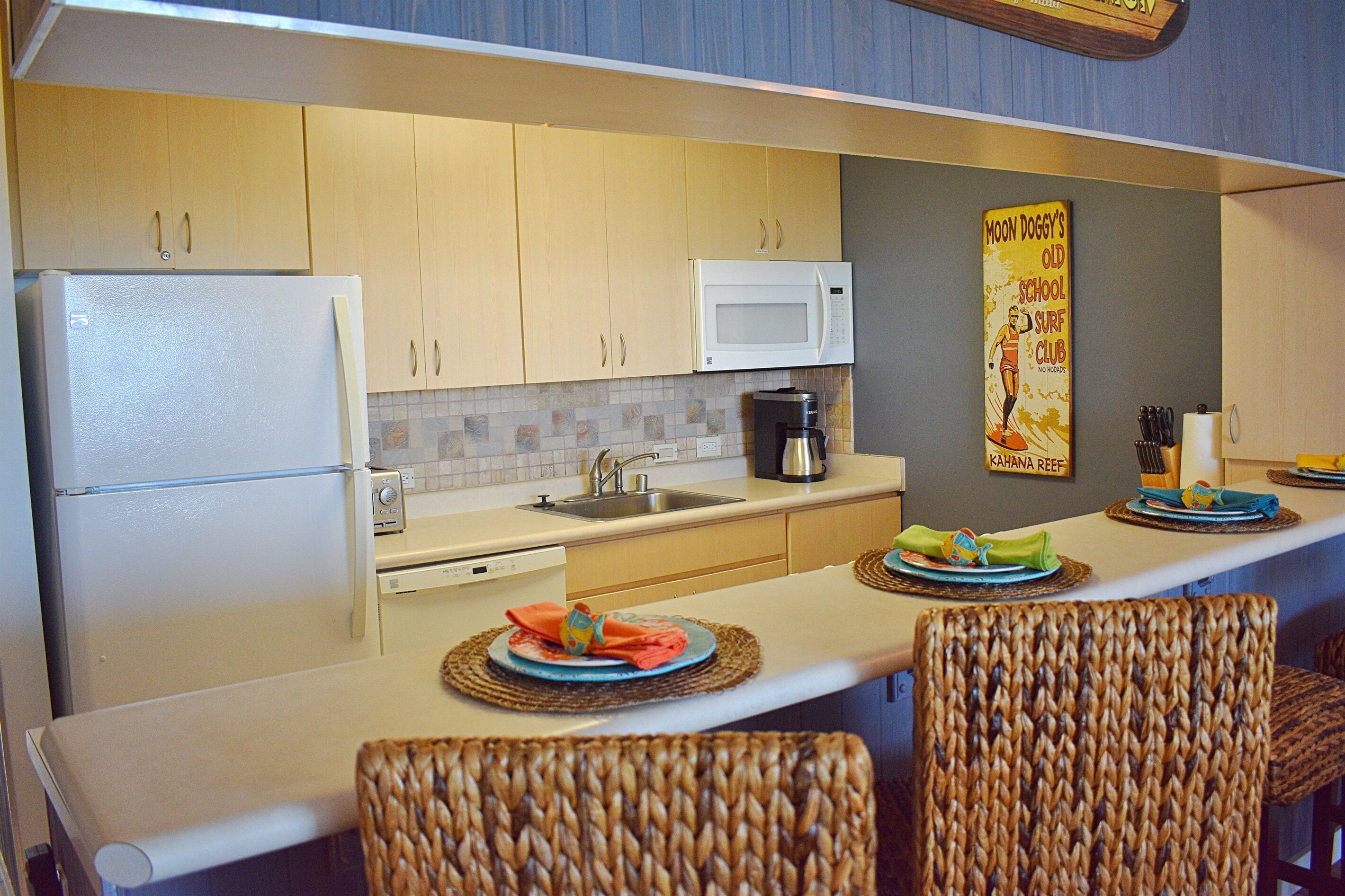 A compact kitchen with a refrigerator, microwave, sink, and a coffee maker. There are wicker chairs and a small table with plates and napkins.