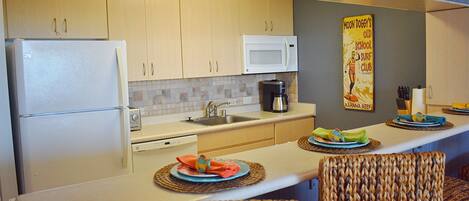 A compact kitchen with a refrigerator, microwave, sink, and a coffee maker. There are wicker chairs and a small table with plates and napkins.