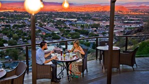 A couple dining at a table with a city view, surrounded by wicker chairs and tables, on a wooden deck. - St. George Gem, Pool, Hot Tub, View, Zion Basecamp (Hurricane)