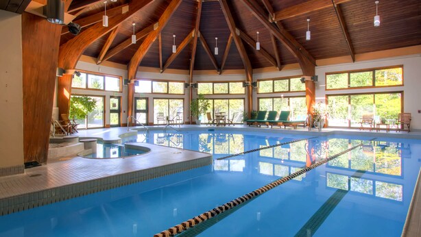 An indoor swimming pool with wooden beams, clear windows, and lounge chairs. - Village 43 | Spacious End Unit with Loft, Fireplace & Resort Amenities (Killington)