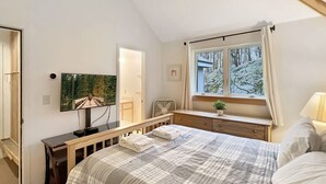 A bedroom with a bed, a desk, a chair, a window with a view of a snowy landscape, and a painting on the wall. - Village 43 | Spacious End Unit with Loft, Fireplace & Resort Amenities (Killington)