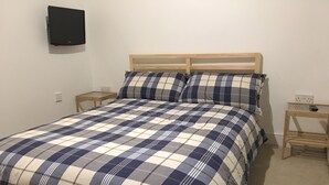 Una cama con colcha escocesa, dos mesitas de noche de madera, un altavoz montado en la pared y un enchufe en la pared.