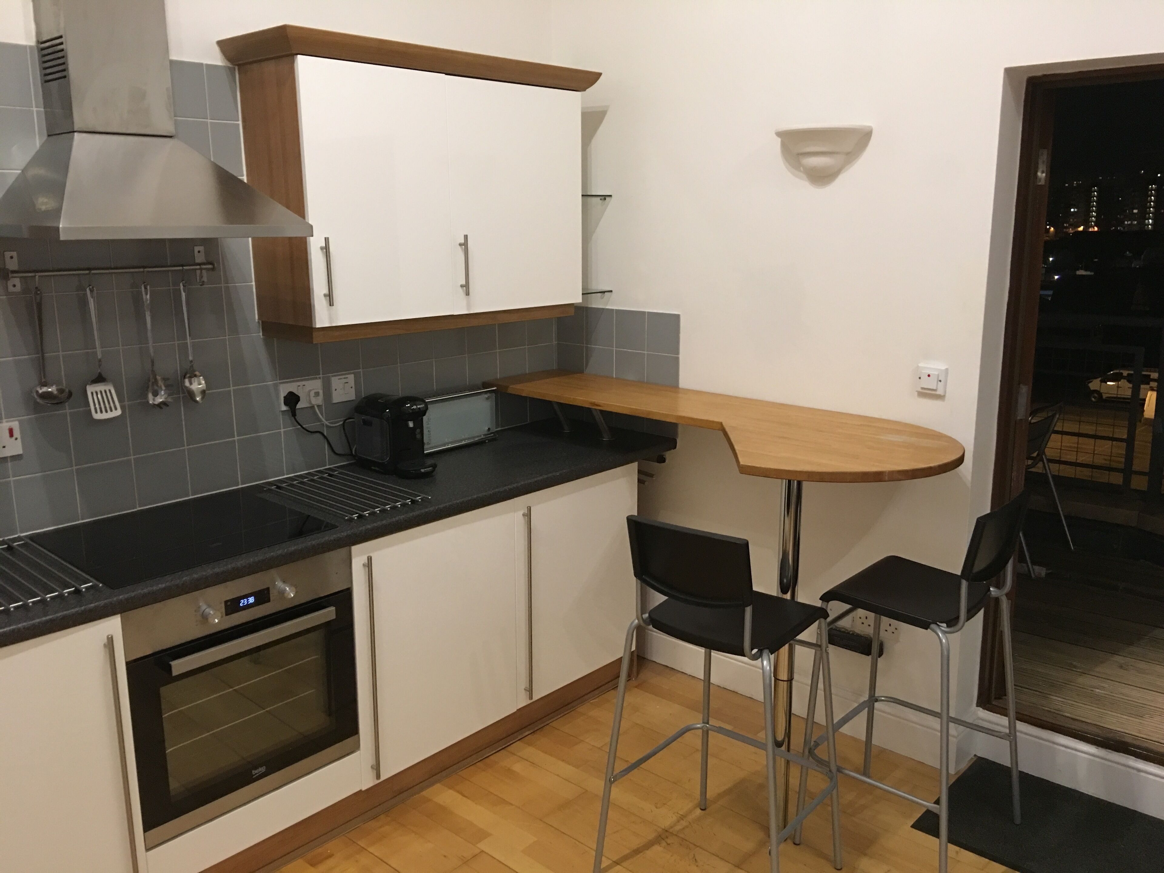 Cocina compacta con horno integrado, cafetera y tostadora. Hay dos taburetes de bar y una mesa de madera pequeña.