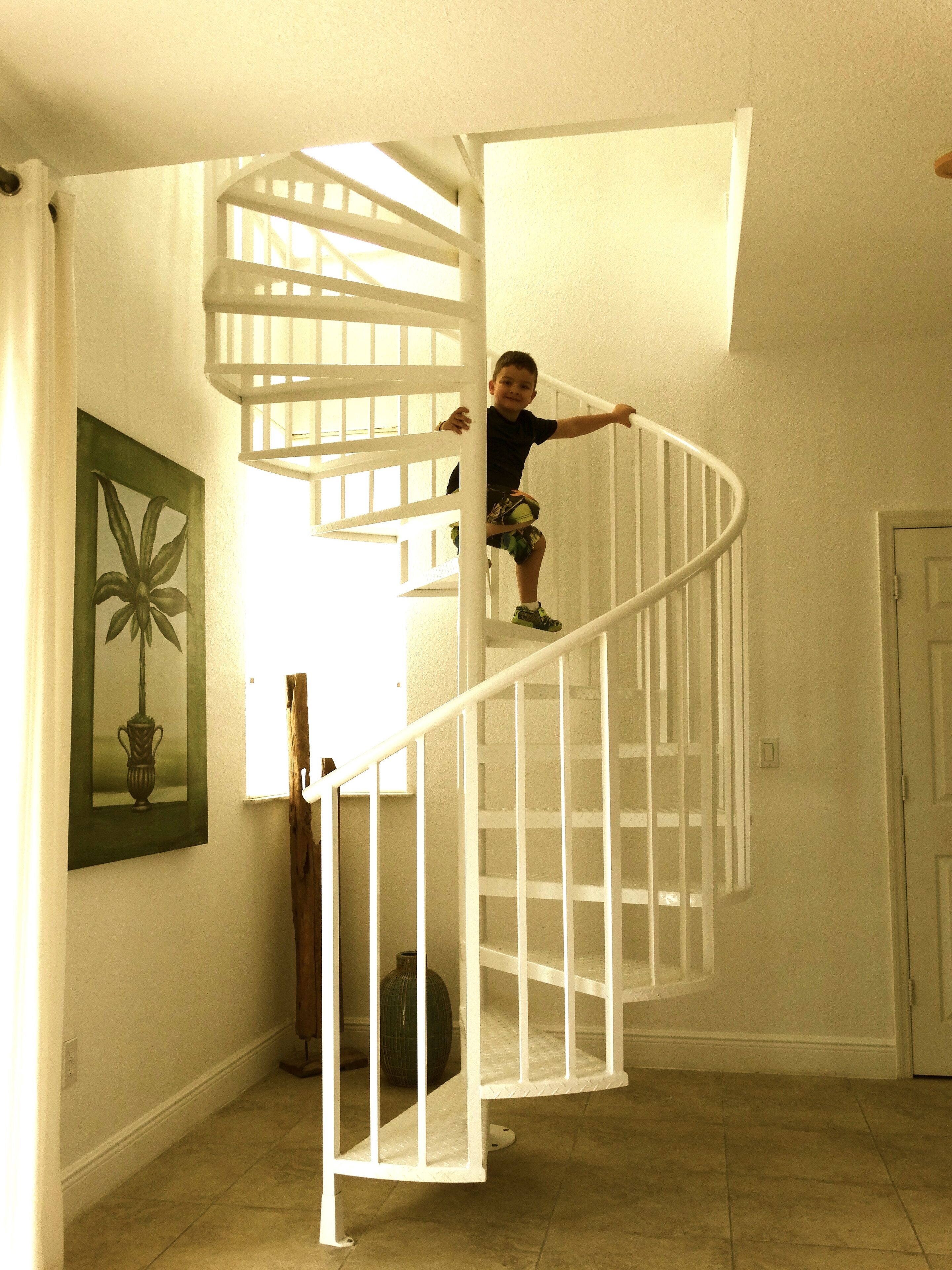 A child climbing a white spiral staircase in a room with a tiled floor, a framed picture on the wall, and a door in the background.