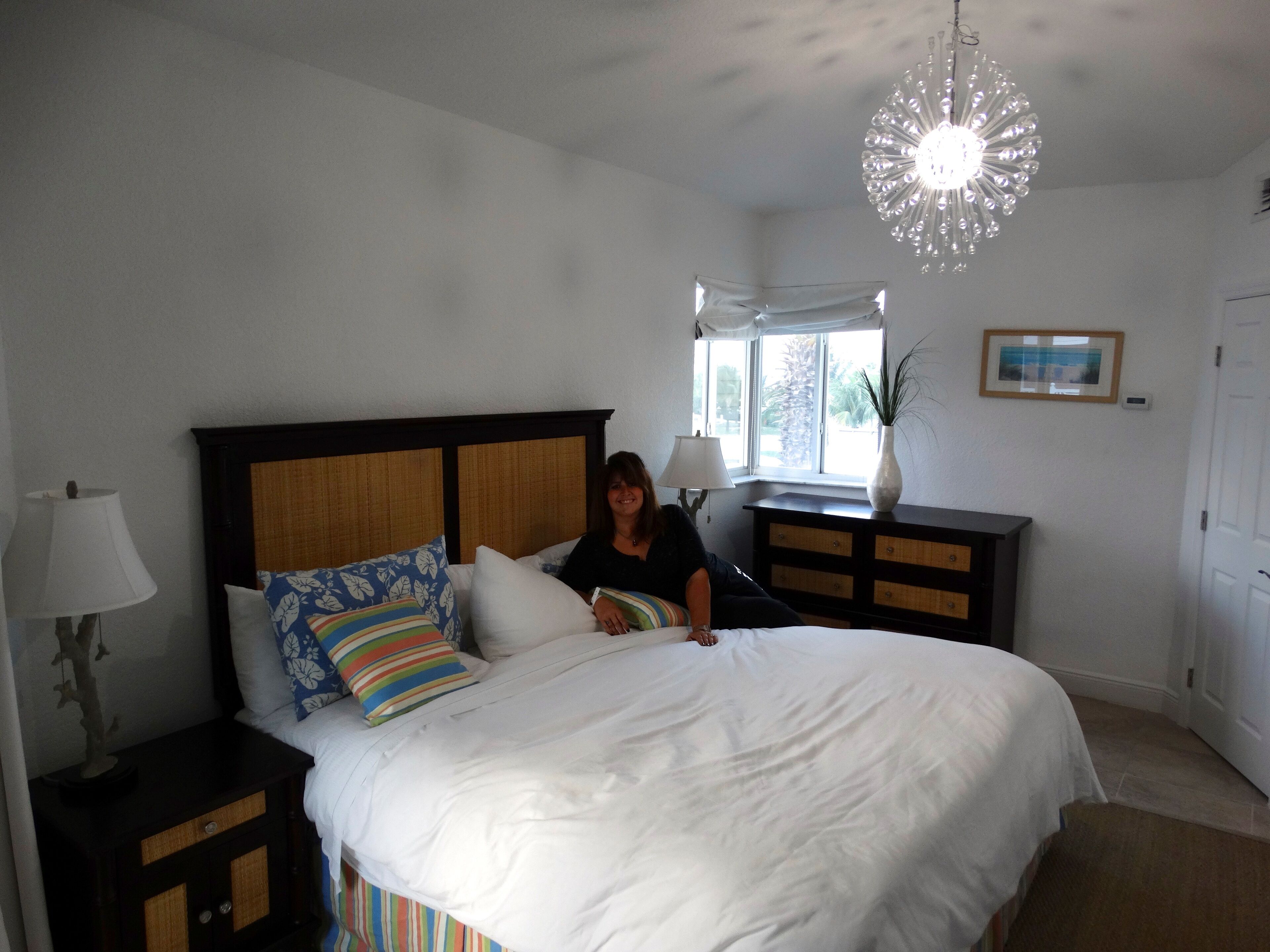 A bedroom with a large bed, a nightstand with a lamp, a dresser, and a window with a view of palm trees.